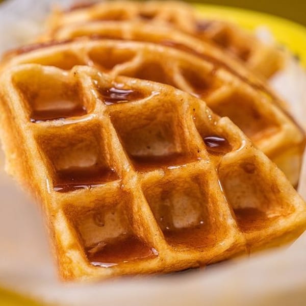 Golden-brown Stack House waffles, drizzled with syrup, in a white basket against a yellow background.