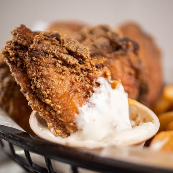 Crispy fried chicken piece being dipped into a small white bowl of creamy sauce.